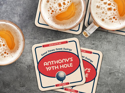 Custom square pulpboard coasters are shown sitting on a dark gray stone background with beer mugs surrounding them. The beer mugs have coasters underneath them. The custom coasters feature a 19th hole bar design with an illustration of a golf ball. The tagline, bar name, established year, and location can be customized on these.
