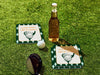 Custom square pulpboard coasters are shown sitting on a grassy, green golf course next to a bottle of beer, white golf ball, and golf club. The coaster features design with an illustration of a golf ball and with club behind it, argyle background pattern, and custom text.