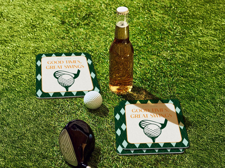 Custom square pulpboard coasters are shown sitting on a grassy, green golf course next to a bottle of beer, white golf ball, and golf club. The coaster features design with an illustration of a golf ball and with club behind it, argyle background pattern, and custom text.