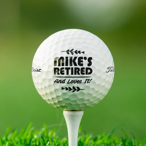 Personalized retirement golf ball featuring the "Retired And Loves It" design with a custom name. Displayed on a golf tee against a golf course background.