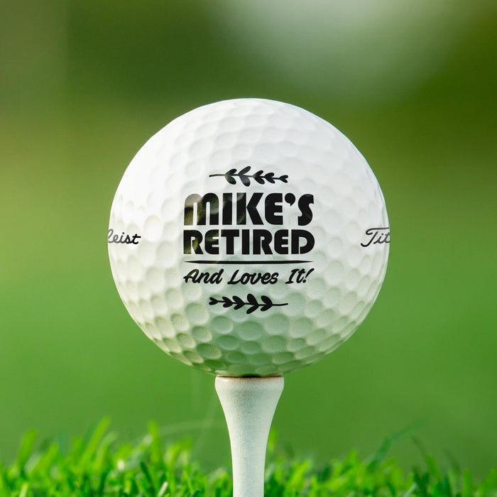 Personalized retirement golf ball featuring the "Retired And Loves It" design with a custom name. Displayed on a golf tee against a golf course background.