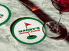 Coaster is shown with a wine glass on top of it and another coaster is off to the side. The custom coasters features a green border, golf ball, a red flag, and golf hole illustration and bold text across the center that can be customized with a name and lounge name.