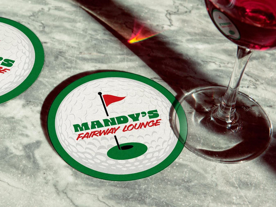Coaster is shown with a wine glass on top of it and another coaster is off to the side. The custom coasters features a green border, golf ball, a red flag, and golf hole illustration and bold text across the center that can be customized with a name and lounge name.