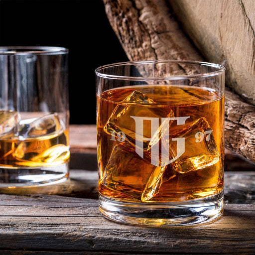 Lifestyle image of a personalized whiskey glass with ice on a wooden table, featuring a bold “AG” monogram with year on each side. The setting includes another glass in the background, creating a warm, bar-inspired atmosphere.