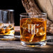 Lifestyle image of a personalized whiskey glass with ice on a wooden table, featuring a bold “AG” monogram with year on each side. The setting includes another glass in the background, creating a warm, bar-inspired atmosphere.