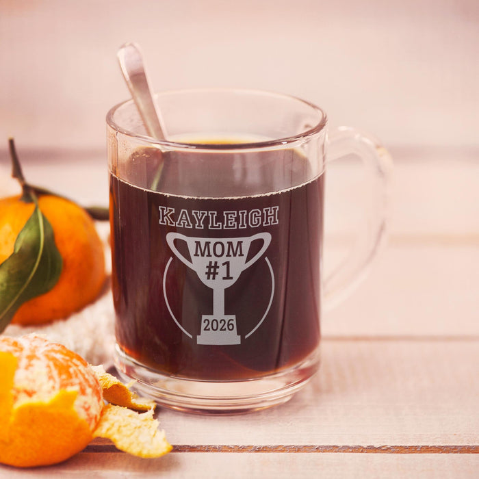 A glass mug filled with a dark liquid, likely coffee, sits on a wooden surface. The mug has a personalized message engraved on it, which reads "Kayleigh Mom #1 2026".