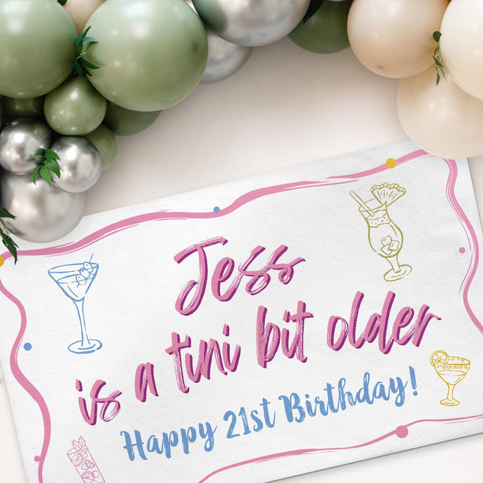 Personalized birthday banner displayed beneath a balloon garland with cocktail-inspired illustrations and custom text.