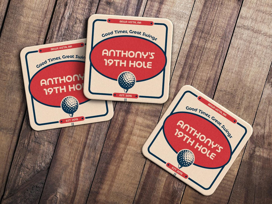 Custom square pulpboard coasters are shown sitting on a wooden table. Three custom coasters feature a 19th hole bar design with an illustration of a golf ball. The tagline, bar name, established year, and location can be customized on these.