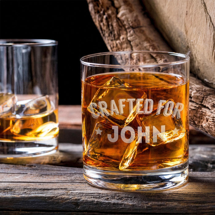 Lifestyle image of the personalized whiskey glass with whiskey and large ice cubes placed on a wooden table beside another drink. The “Crafted For John” engraving gives the glass a refined and personalized bar-inspired look.