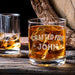 Lifestyle image of the personalized whiskey glass with whiskey and large ice cubes placed on a wooden table beside another drink. The “Crafted For John” engraving gives the glass a refined and personalized bar-inspired look.