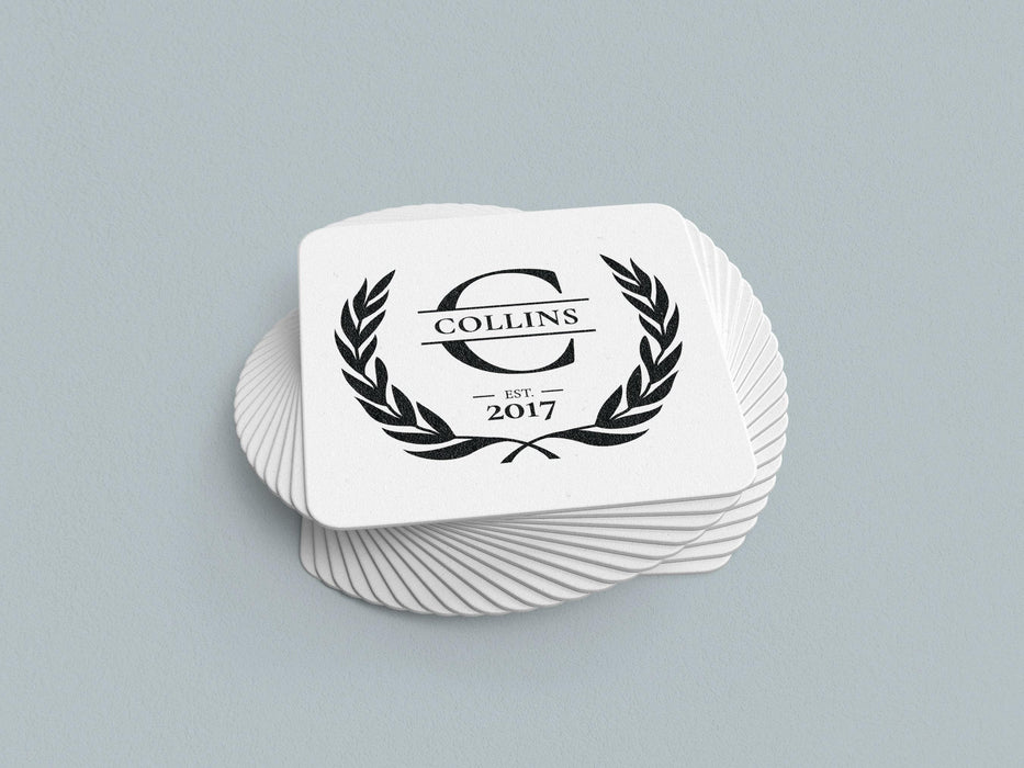 Laurel Monogram design shown on square paper coasters sitting on a blue gray surface.