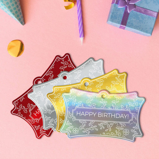 Four floral birthday hang tags in metallic gold, metallic silver, metallic red, and metallic rainbow lying beside colorful gifts and party decorations.