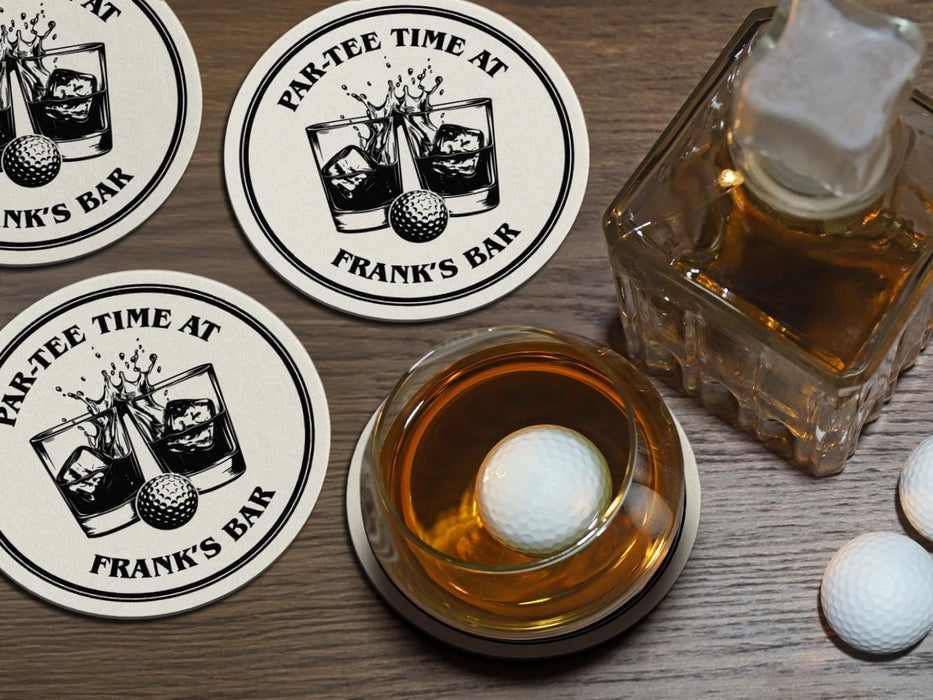 A full bourbon glass is shown next to a decanter while sitting on a wooden table. A golf ball rests in the glass. Custom coasters and golf balls can be seen on the table as well. A custom coaster is also placed underneath the glass. The custom coasters feature a golf ball centered between two whiskey glasses clinking together and splashing out with customizable text wrapping around the illustration. The coaster design is printed in black.