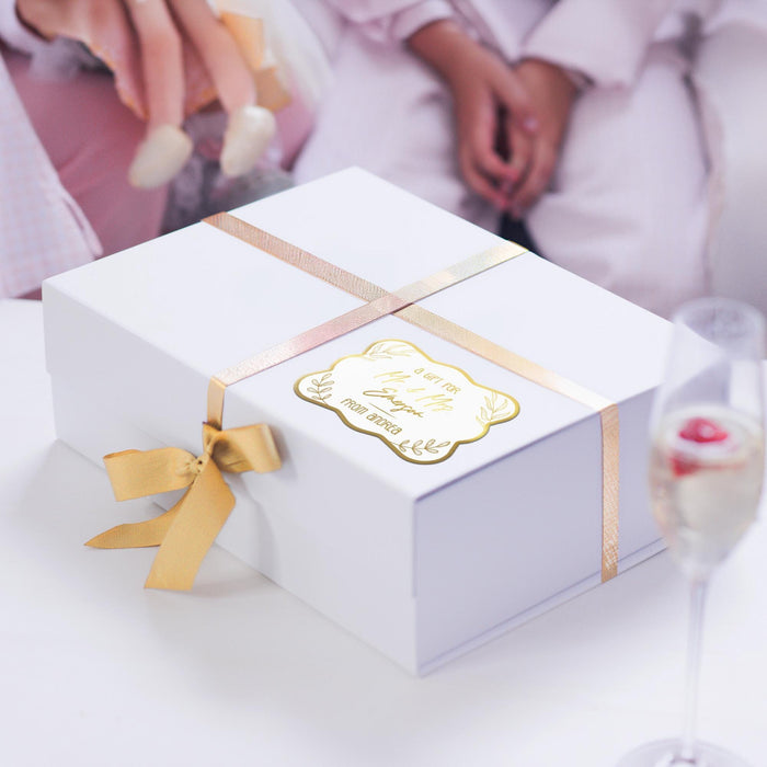 White gift box wrapped with satin ribbon and decorated using a personalized “Mr. & Mrs.” gold foil wedding gift tag. Styled as premium wedding and bridal shower gift packaging.
