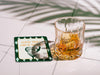Custom square pulpboard coaster are shown laid out on marble floor. A whiskey glass is shown next to the coaster. The coaster features design with an illustration of a golf ball and with club behind it, argyle background pattern, and custom text.