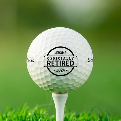 Single white Titleist golf ball with Officially Retired design on white golf tee with golf course grass in the background
