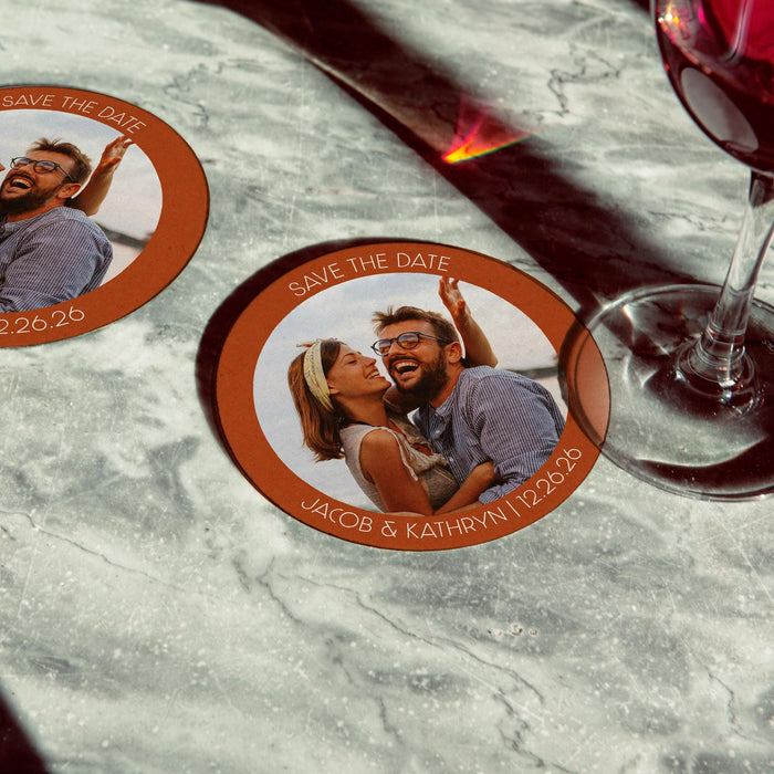 Personalized photo Save the Date coaster displayed beside a wine glass. Custom wedding favor featuring a couple photo and wedding details.