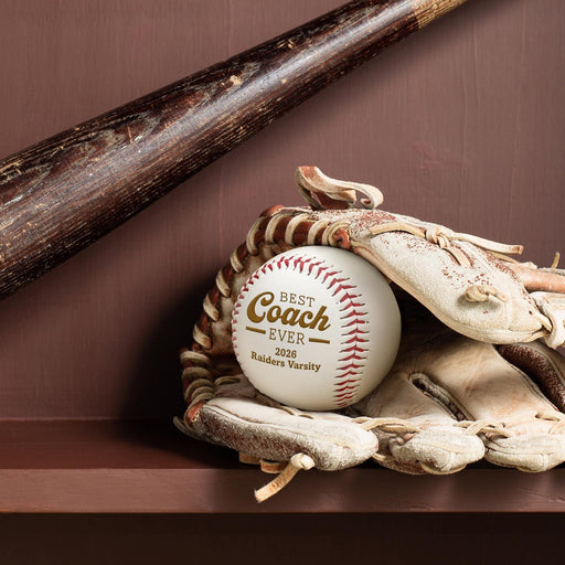 A baseball bat and a baseball glove are placed on a shelf, with a personalized baseball featuring the text "Best Coach Ever", the year "2026" and a team/school name "Raiders Varsity" engraved on it.