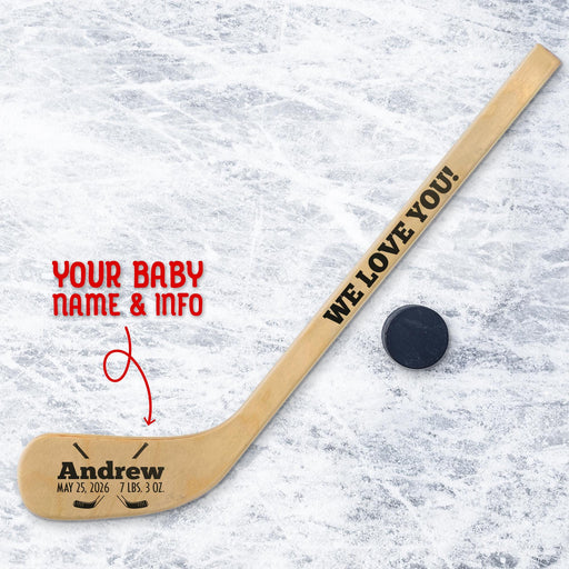 Personalized mini wooden hockey stick featuring baby name and birth details in a clean, centered layout with a bold name and smaller birth stats beneath, accented by a hockey puck graphic, shown on an ice background with a real puck.