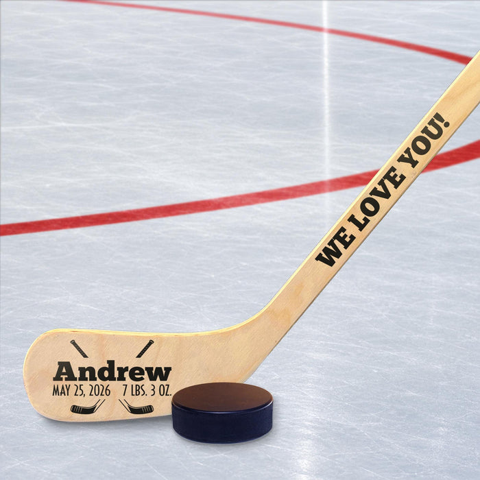 Personalized hockey stick displayed on an ice rink with baby name and birth details printed in a bold layout with hockey-themed graphics, placed next to a real puck.