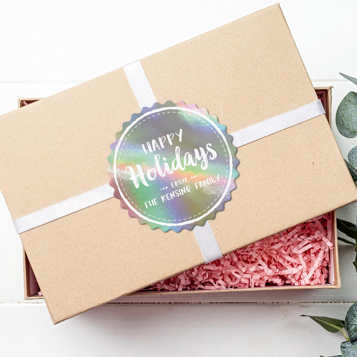 Personalized round Happy Holidays gift tag displayed on a kraft gift box with white ribbon and pink crinkle paper.