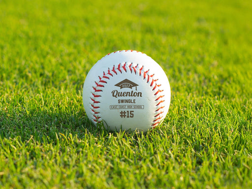 A custom baseball sits outside on green grass. This baseball features an engraved design that uses a graduation cap graphic with the player's graduating year, their first and last name, school name, and jersey number.