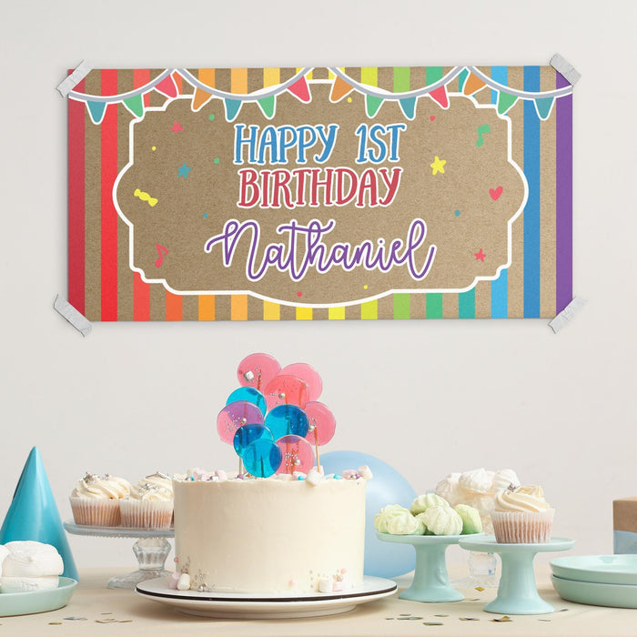 First birthday dessert table setup featuring a custom rainbow birthday banner displayed above a birthday cake and party decorations.