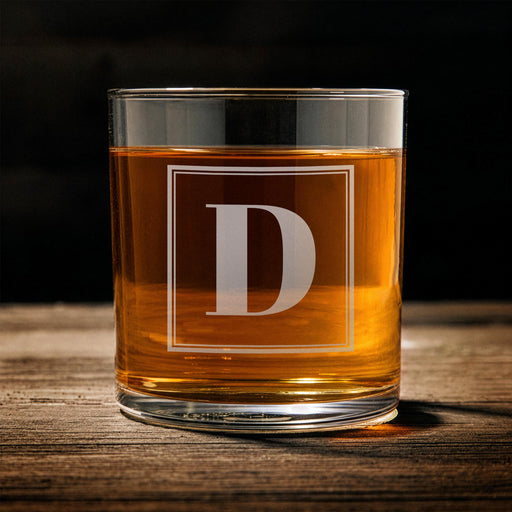 Front-facing product image of a personalized whiskey glass on a wooden table, featuring a framed “D” initial design. The clean layout highlights the simple and elegant engraving style.
