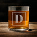 Front-facing product image of a personalized whiskey glass on a wooden table, featuring a framed “D” initial design. The clean layout highlights the simple and elegant engraving style.