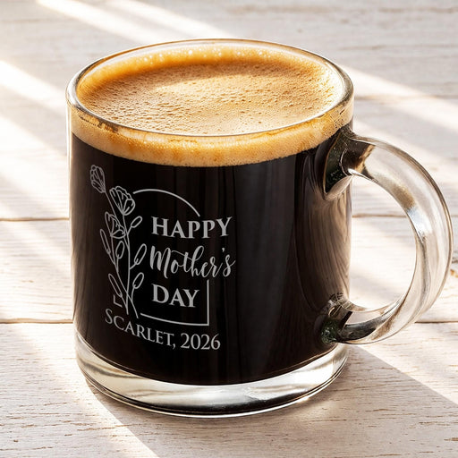 A glass mug filled with a dark liquid, likely coffee, with a frothy top. The mug has an engraved message that reads "Happy Mother's Day SCARLET, 2026".