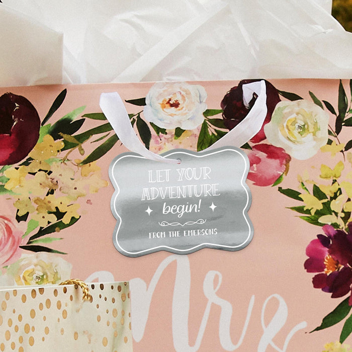 Romantic floral flat lay featuring a personalized “Let Your Adventure Begin” wedding gift tag in metallic silver foil. Styled with flowers and wedding-inspired décor.