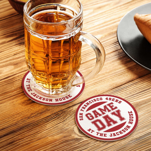 Red custom football coaster under a beer mug with another personalized Game Day coaster displayed beside it on a wooden table.
