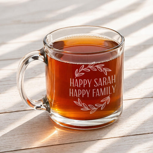 A glass mug filled with a warm beverage, likely tea, sits on a wooden surface. The mug has a personalized message engraved on it that reads "Happy Sarah Happy Family".