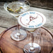 Personalized martini coaster displayed beside two martini glasses on a wooden table, featuring the custom bar name, martini illustration, and established year.