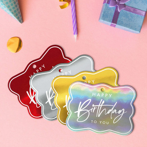 Four elegant script hang tags in metallic gold, metallic silver, metallic red, and metallic rainbow lying beside colorful gifts and party decorations.