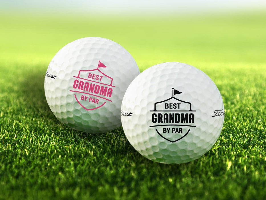 Two golf balls with the text "BEST GRANDMA BY PAR" printed on them, placed on a grassy field.