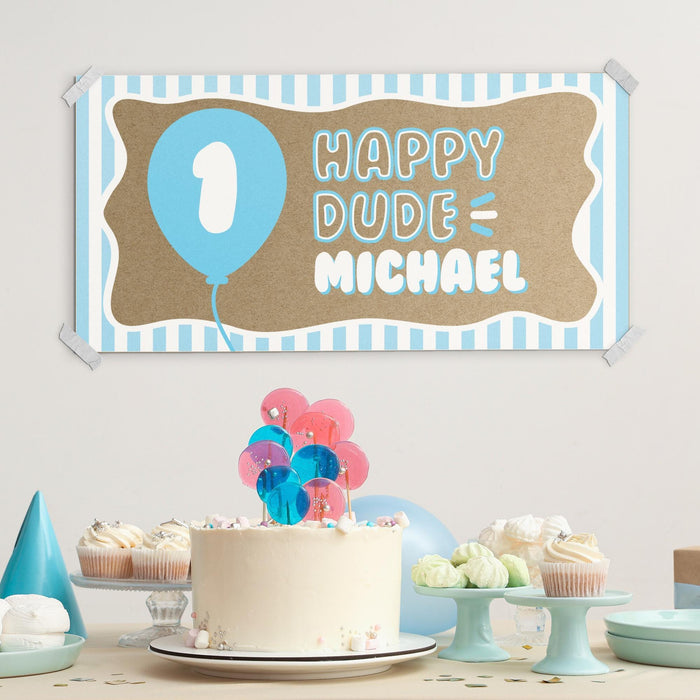 First birthday party setup featuring a custom printed Happy Dude banner displayed above a dessert table with birthday cake and decorations.