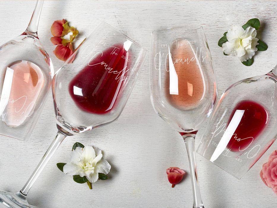 Four wine glasses lay on white wooden surface next to flowers. The glasses have Last Name and Established Date design engraved on it.