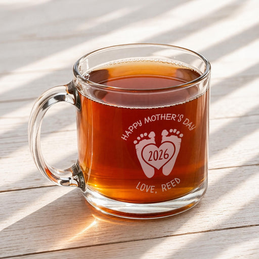 A glass mug filled with a warm beverage, likely tea, sits on a wooden surface. The mug has a personalized message engraved on it, celebrating Mother's Day with the year 2026 and the names 'Love, Reed'.