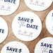 Personalized Save the Date coasters shown in a wedding favor arrangement. Features a modern typography design with custom names and wedding date.