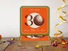 Custom square pulpboard coasters are shown on a wooden surface with confetti surrounding them. The coaster features design with illustrated golf ball, custom age and year, and vivid orange, cream, and green background.