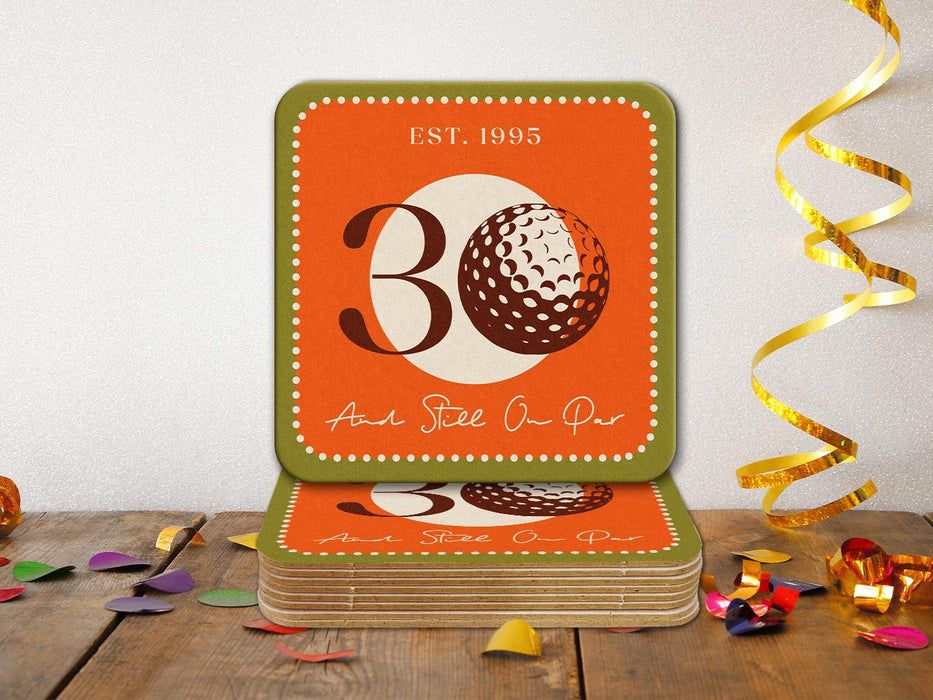 Custom square pulpboard coasters are shown on a wooden surface with confetti surrounding them. The coaster features design with illustrated golf ball, custom age and year, and vivid orange, cream, and green background.