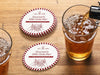 Custom round pulpboard coasters are shown sitting on a wooden table next to two drink glasses. The custom coasters feature a golf club lounge design with an illustration of a golf ball on a tee and people cheersing drinks together along with a striped border. The established year, name, lounge name, and tagline can be customized.