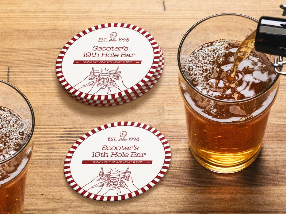 Custom round pulpboard coasters are shown sitting on a wooden table next to two drink glasses. The custom coasters feature a golf club lounge design with an illustration of a golf ball on a tee and people cheersing drinks together along with a striped border. The established year, name, lounge name, and tagline can be customized.