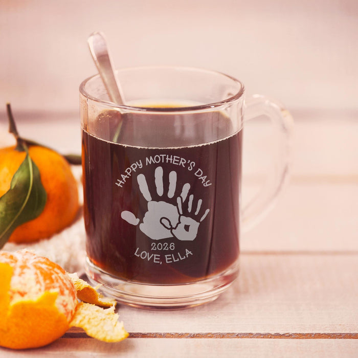 A glass mug filled with a dark liquid, likely coffee, sits on a wooden surface. The mug has a handprint design and the text "Happy Mother's Day 2026 LOVE ELLA" engraved on it.