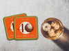Two coasters sit on stone table with whiskey glass next to them. The coaster features design with illustrated golf ball, custom age and year, and vivid orange, cream, and green background.