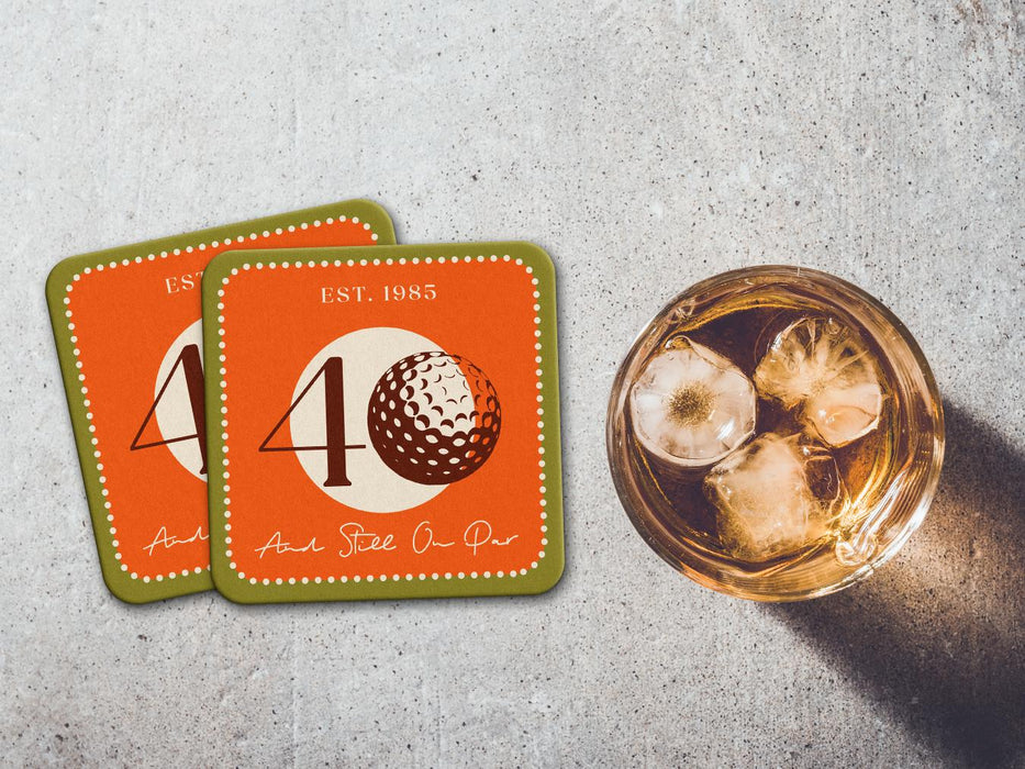 Two coasters sit on stone table with whiskey glass next to them. The coaster features design with illustrated golf ball, custom age and year, and vivid orange, cream, and green background.