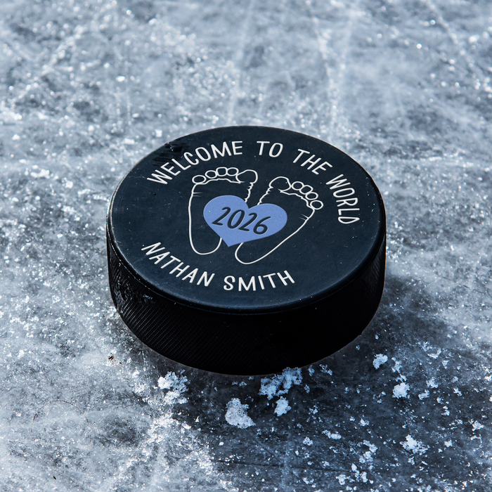 Personalized black hockey puck with a blue UV printed “Welcome to the World” birth announcement design featuring baby feet, a heart, year, and name.
