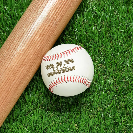 Baseball sits on grass next to baseball bat. Baseball has Established Date Father's Day design engraved on it.