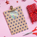Personalized Happy Holidays round gift tag clipped to a polka dot kraft gift bag on a pink holiday background with wrapped presents and scissors.